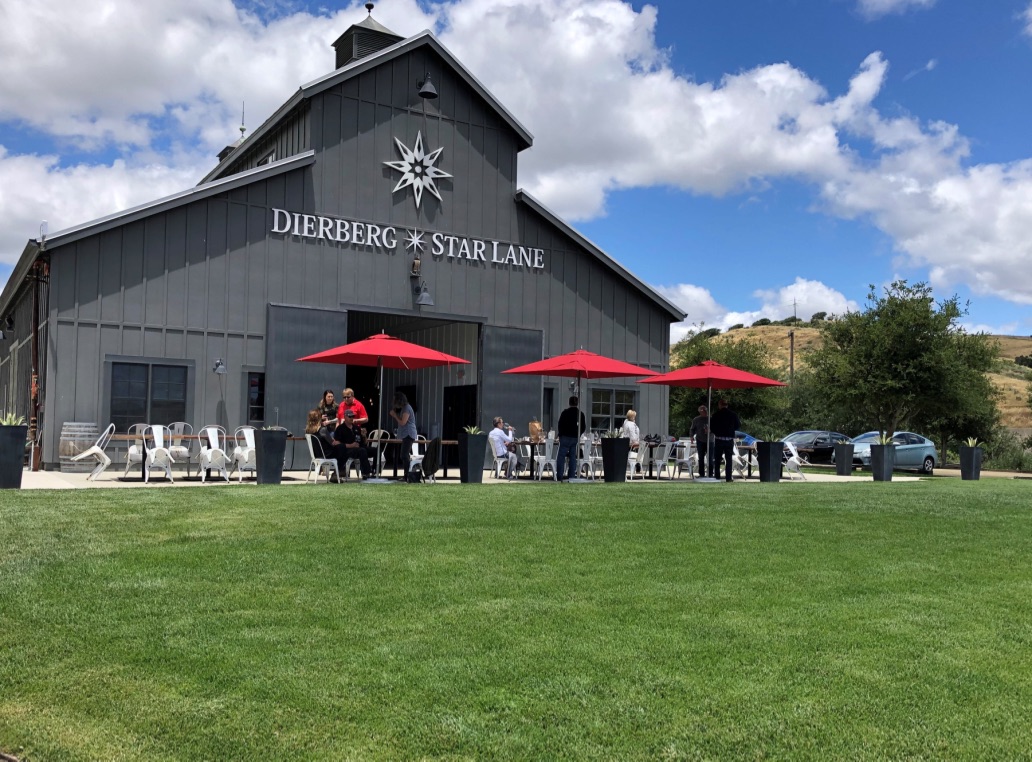 Gray barn-style building with sign, red umbrellas outside, people sitting and standing, green lawn, blue sky with clouds.