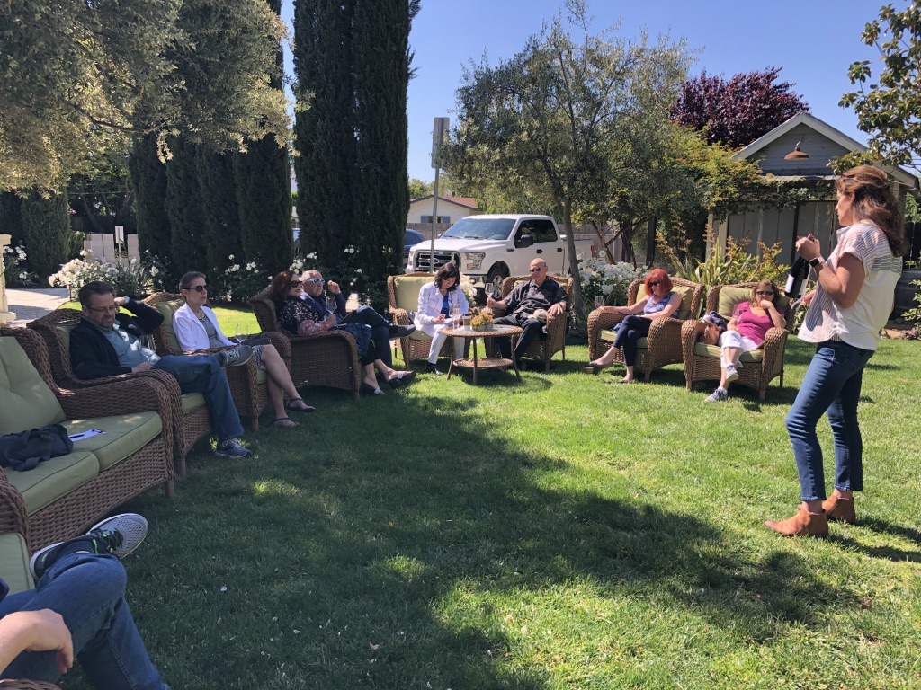Group of people sitting on couches outdoors in a garden, listening to a woman speaking, with trees and houses in background.