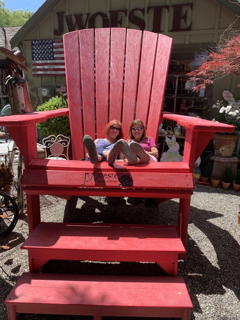 Two children sit on a large red Adirondack chair outdoors, with a building and American flag in background.