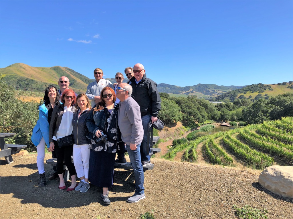 Group of ten people standing outdoors in a vineyard with hills and blue sky in the background.