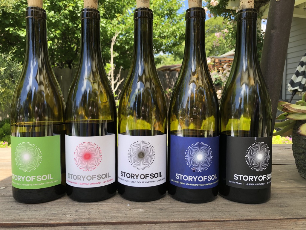 Five wine bottles with different colored labels on a wooden surface outdoors, trees and sky in background.