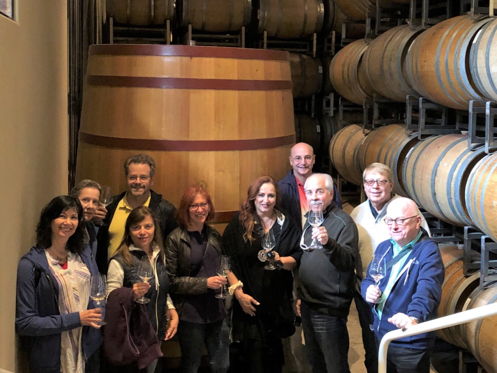 Group of nine people standing in a winery with large wooden barrels in the background, holding glasses.