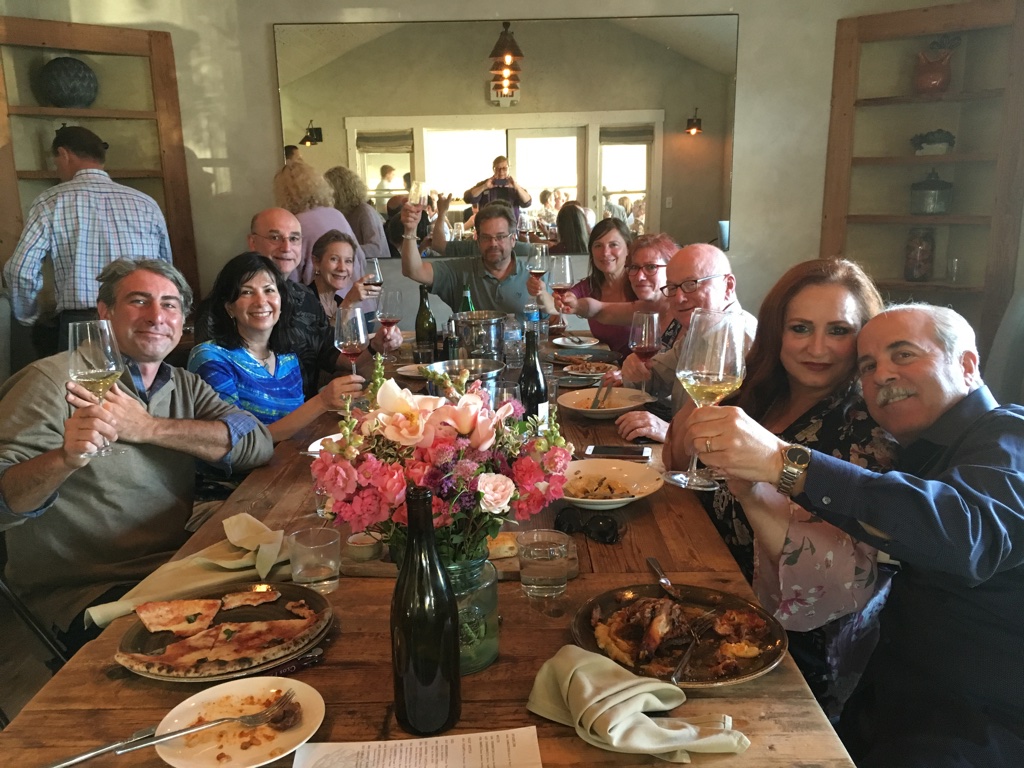 Group of people sitting around a table with food and drinks, celebrating indoors with a flower centerpiece.