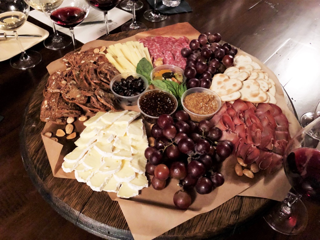 Cheese, grapes, cured meats, and olives on a round wooden platter with wine glasses in background.