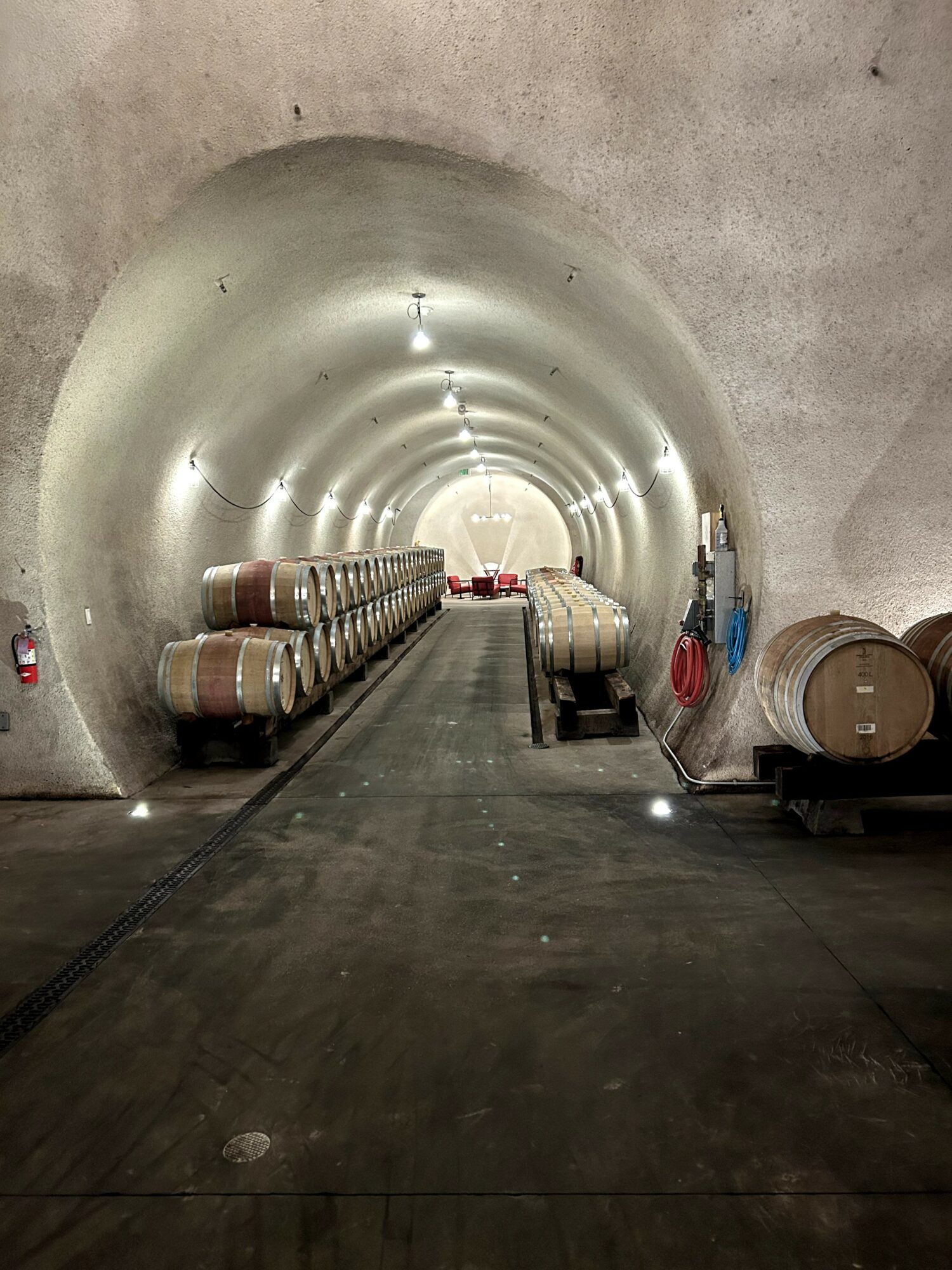Underground tunnel with barrels stacked on sides, concrete walls, and a pathway leading into the distance.