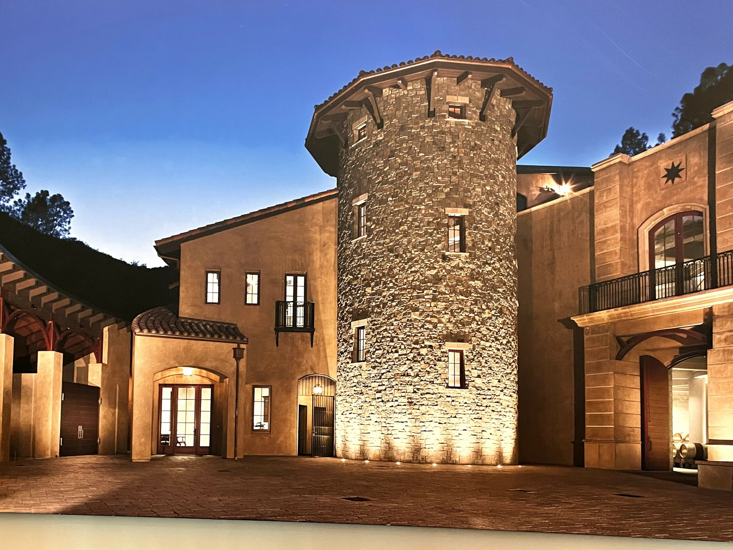 Stone tower with surrounding buildings, illuminated at dusk, with trees in the background.
