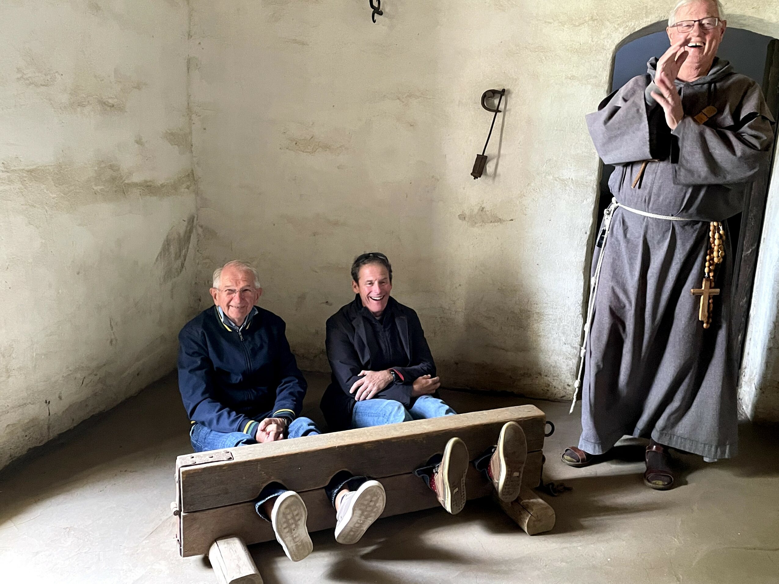 Three men inside a plain room with white walls; two seated on a bench, one standing in a robe with hands clasped.