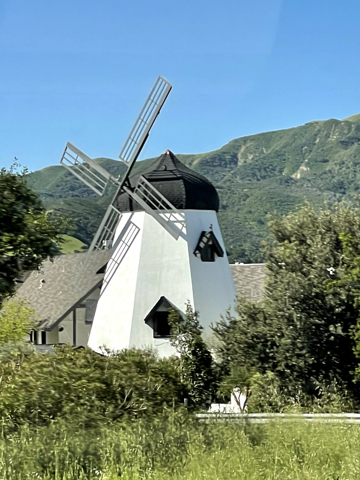 White windmill with black roof and blades, surrounded by greenery and mountains in the background.