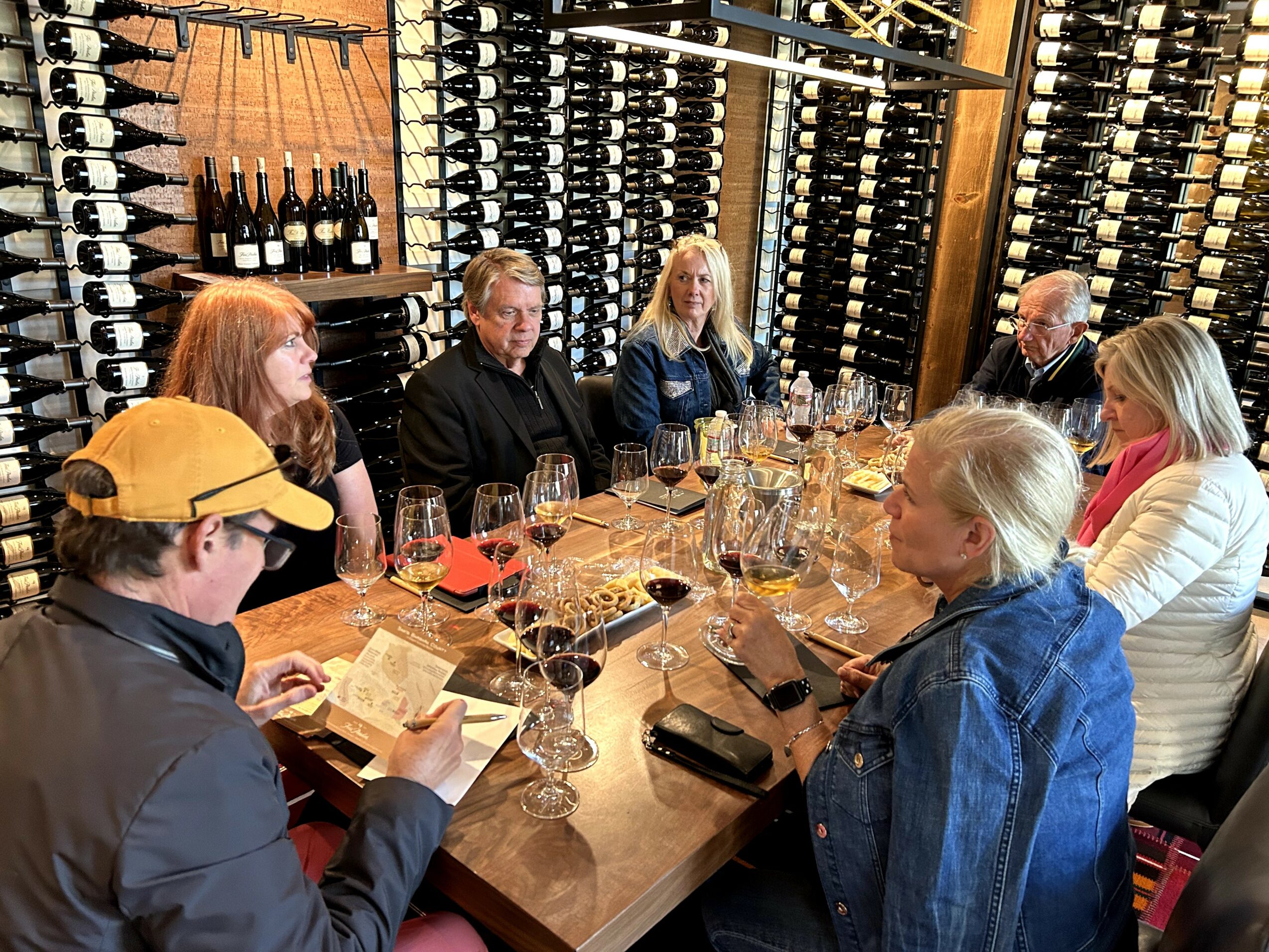 Group of eight people sitting around a wine-tasting table with wine bottles on shelves behind them.