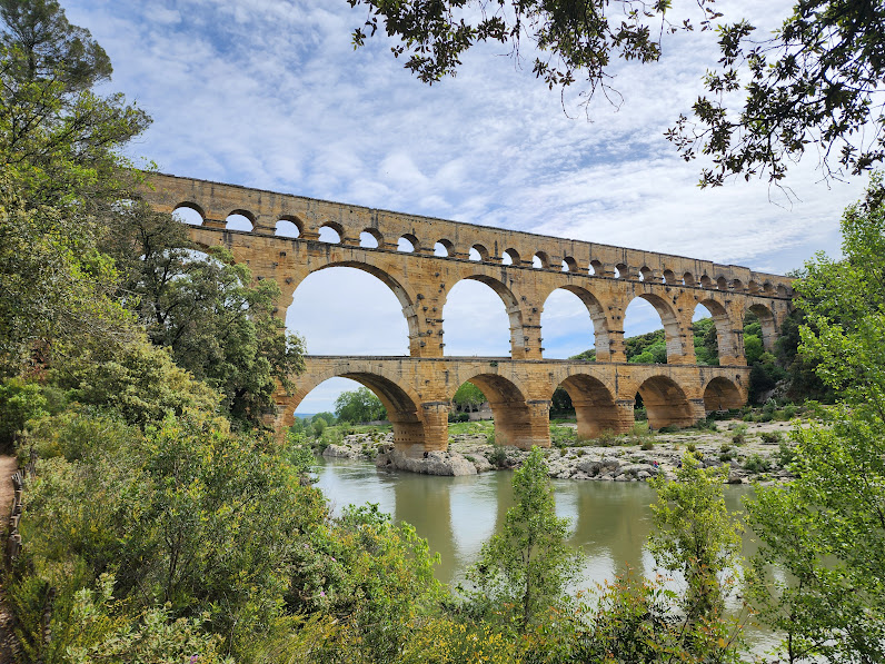 Ancient stone aqueduct with multiple arches spans over a river, surrounded by green trees and a partly cloudy sky.