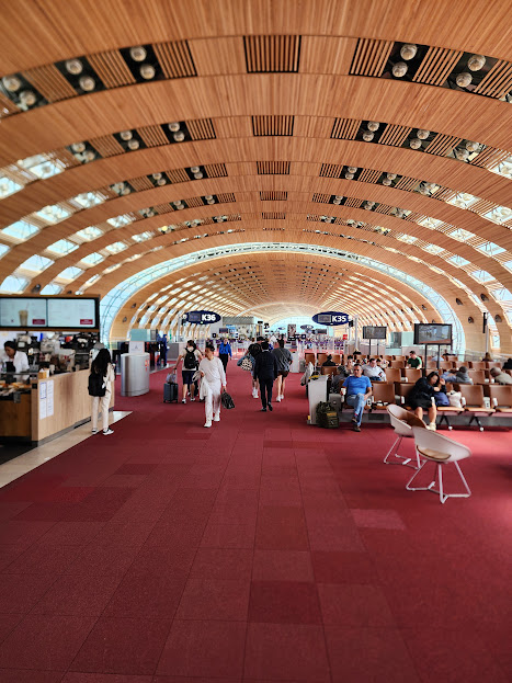 Interior of an airport with people walking and sitting, wooden arched ceiling, digital screens, and check-in counters.