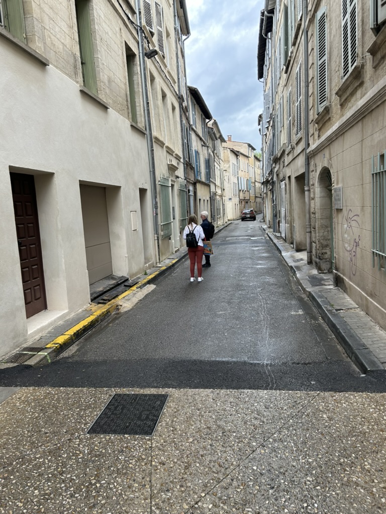Narrow street with buildings on both sides, three people walking in the distance, cloudy sky above.