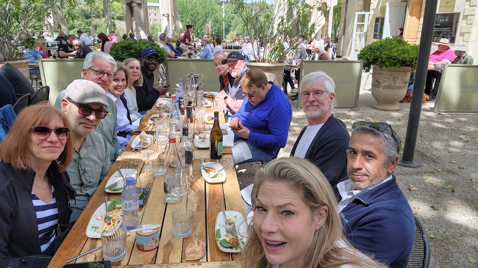Group of people sitting at a long outdoor table with food and drinks, smiling and socializing.