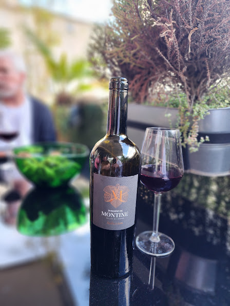 Bottle of red wine and a glass filled with red wine on a reflective table, with blurred background of plants and a person.
