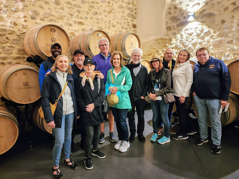 Group of ten people standing in a cellar with large wine barrels, smiling and posing for the photo.