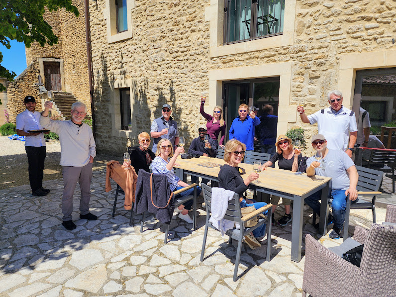 Group of people sitting and standing outside on a stone patio near a stone building, some waving and smiling.