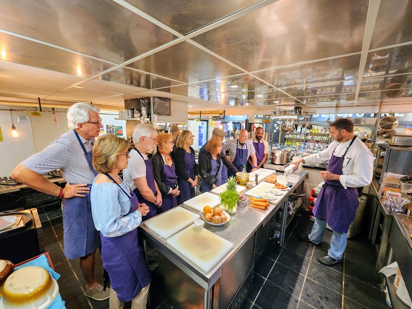 Group of people gathered around a kitchen counter, listening to a chef demonstrating cooking techniques.