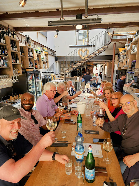 People sitting at a long table in a restaurant, holding wine glasses, with shelves and staircase in background.