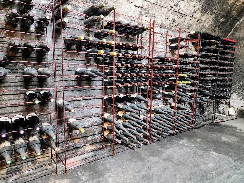 Multiple wine bottles stored horizontally on metal racks against a wall, with some bottles stacked on the ground.
