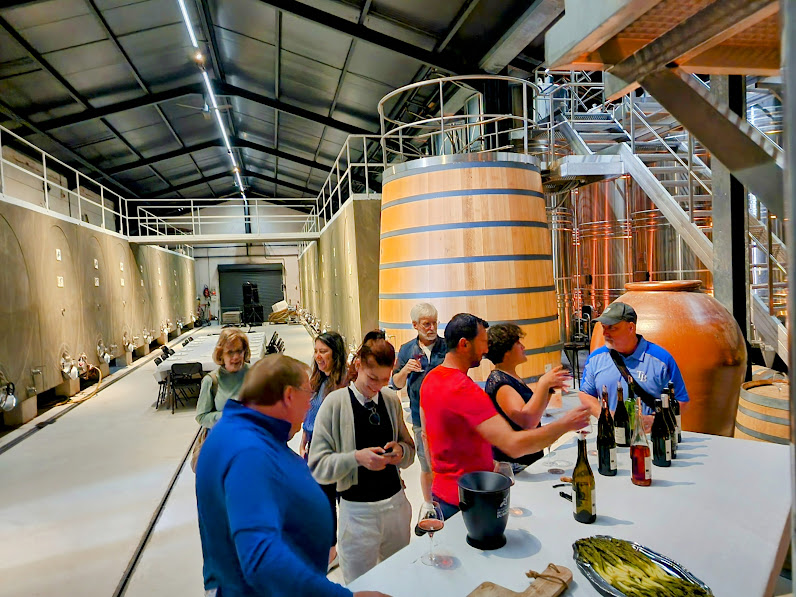 People gather around a white table with bottles and a bowl in an industrial setting with large tanks and metal structures.