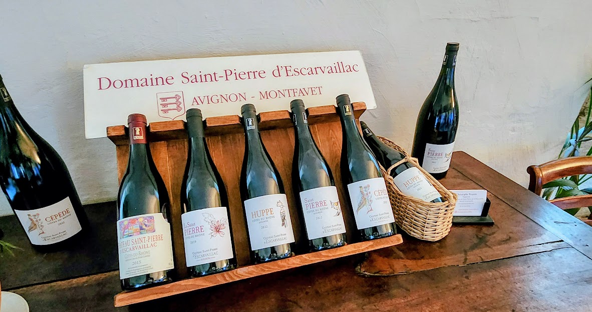 Six wine bottles on a wooden table with a basket and a sign in the background.