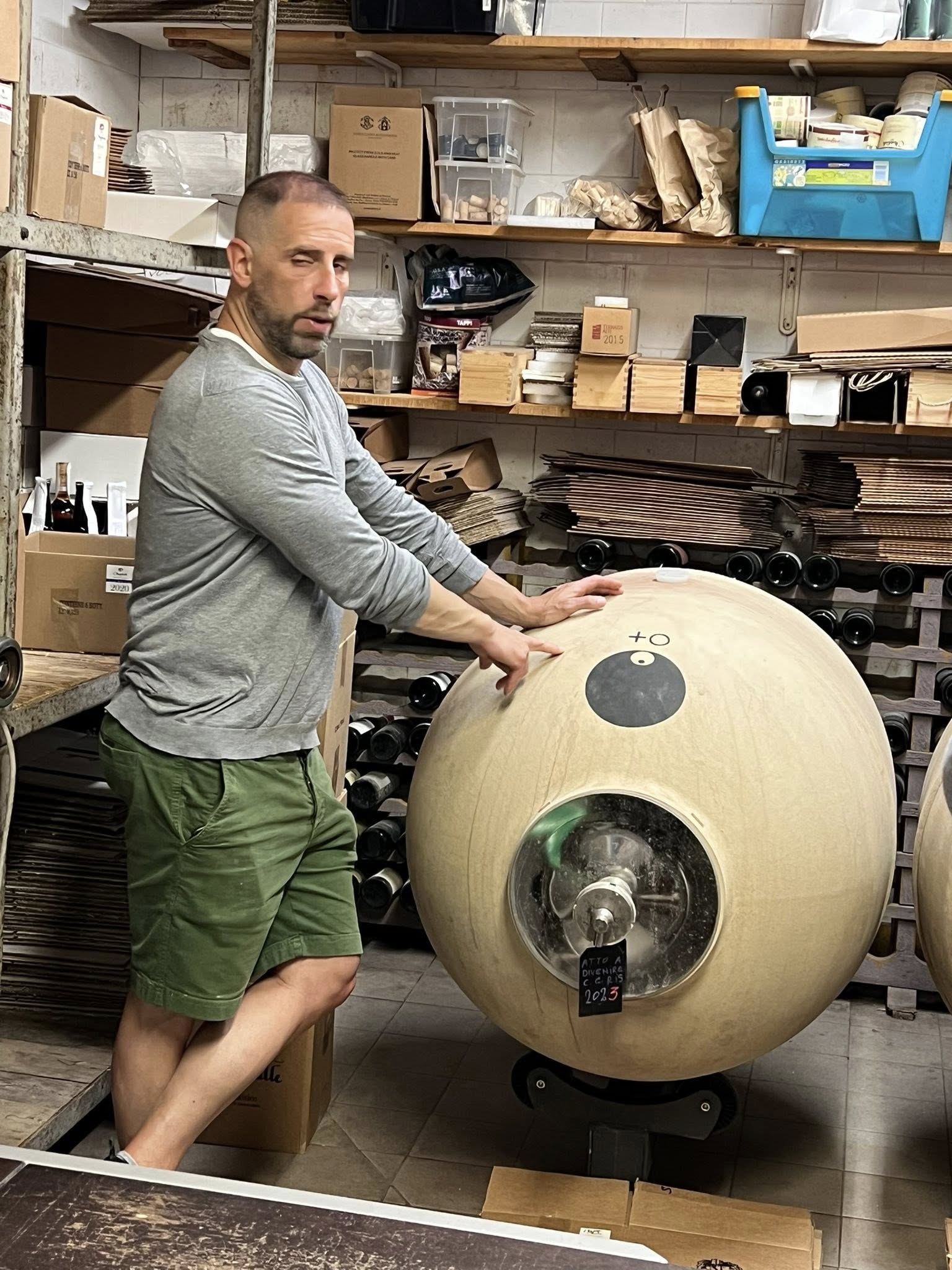 Man in gray shirt and green shorts standing next to a large spherical object in a workshop.