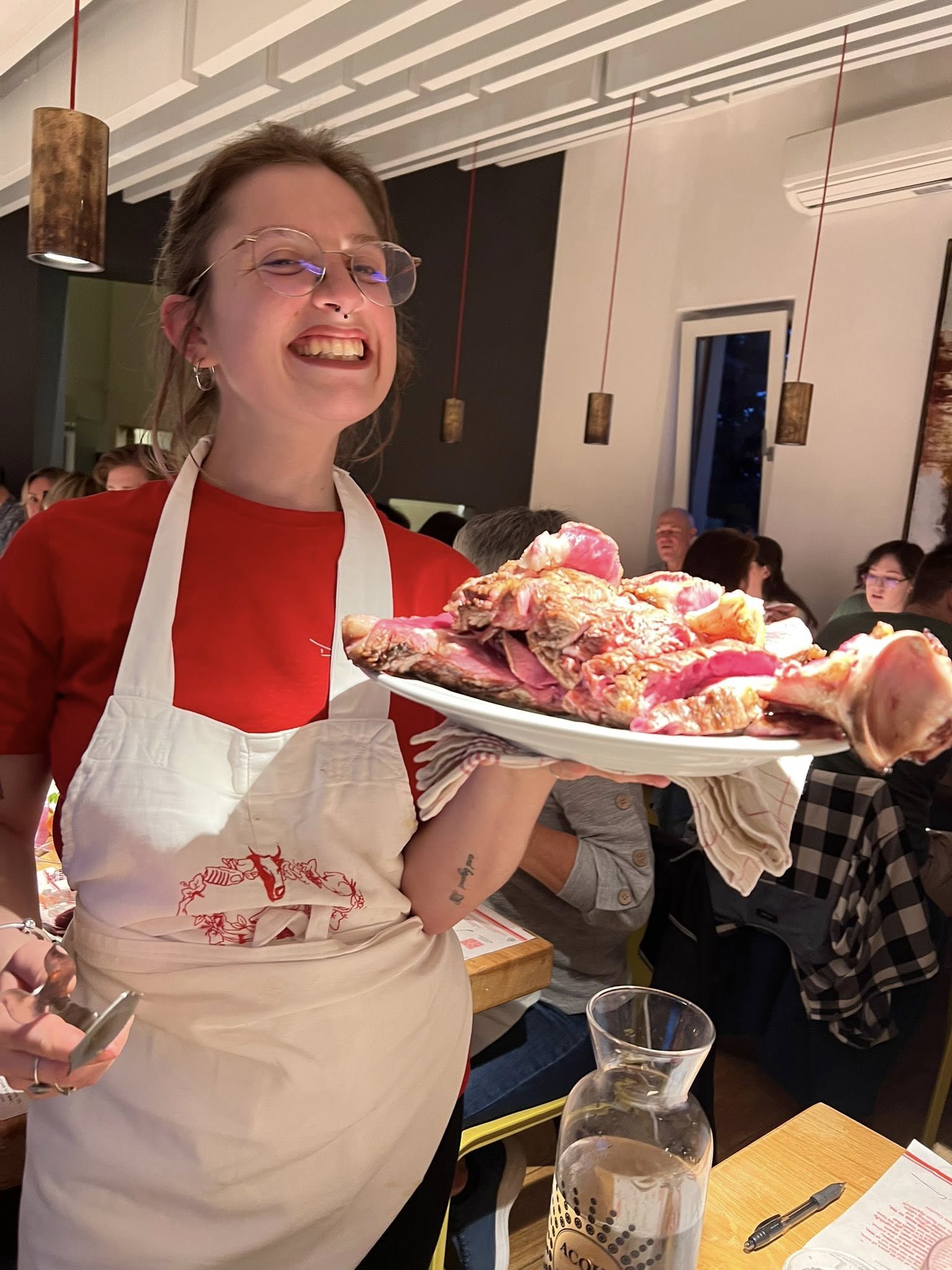 Woman smiling, wearing glasses and apron, holding a plate of raw meat in a restaurant setting.