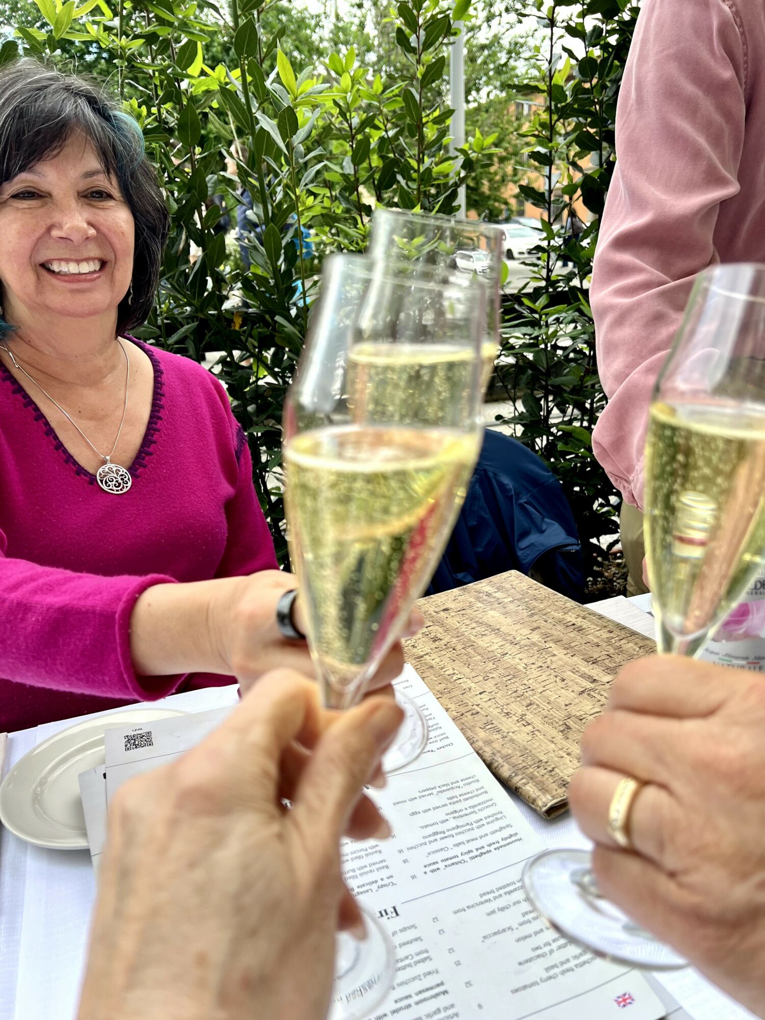 Woman in pink sweater smiling, holding a glass of champagne, with greenery in background, and two other glasses in foreground.