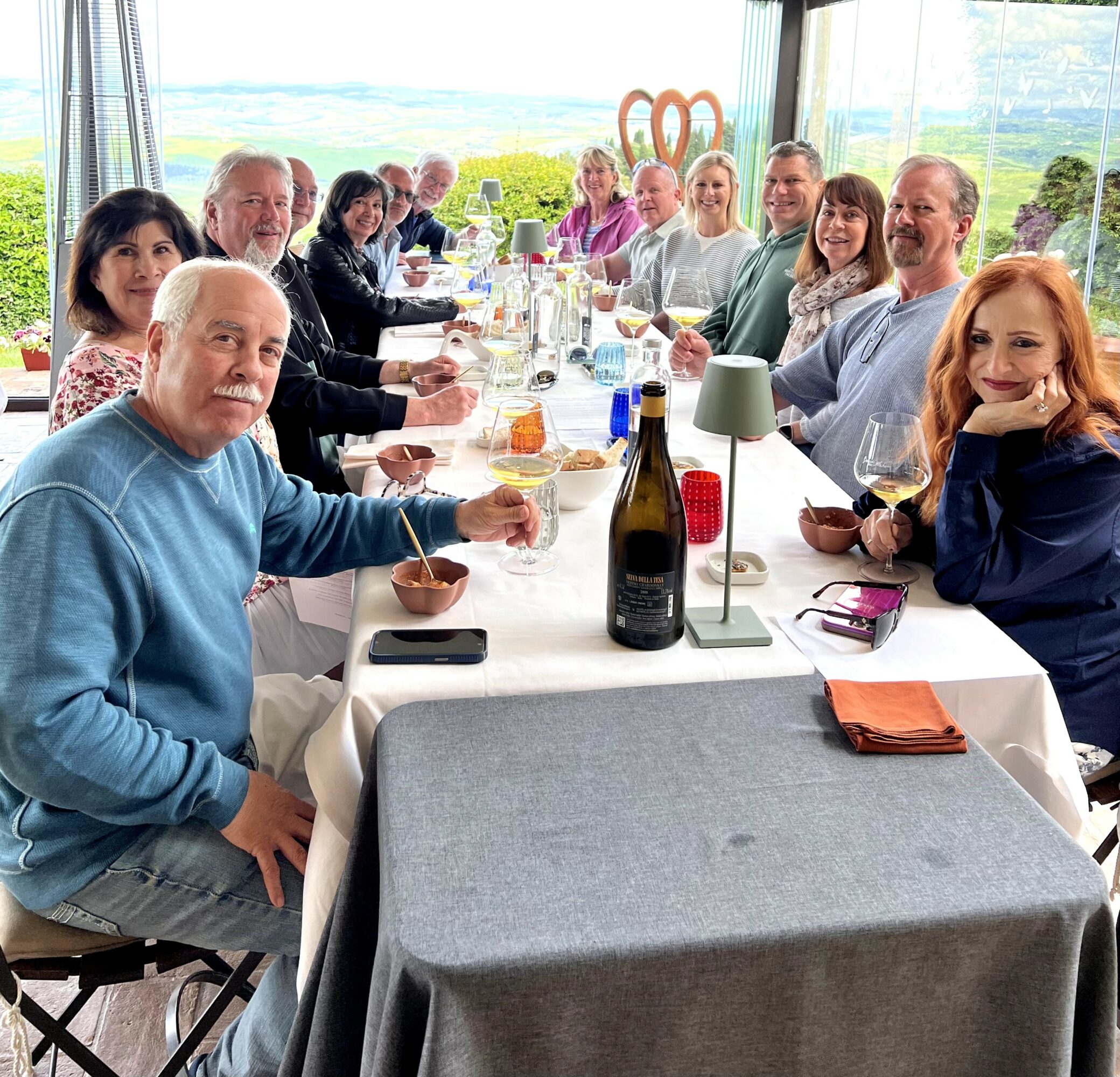 Group of people sitting around a long table with food and drinks, indoors with large windows and a view outside.