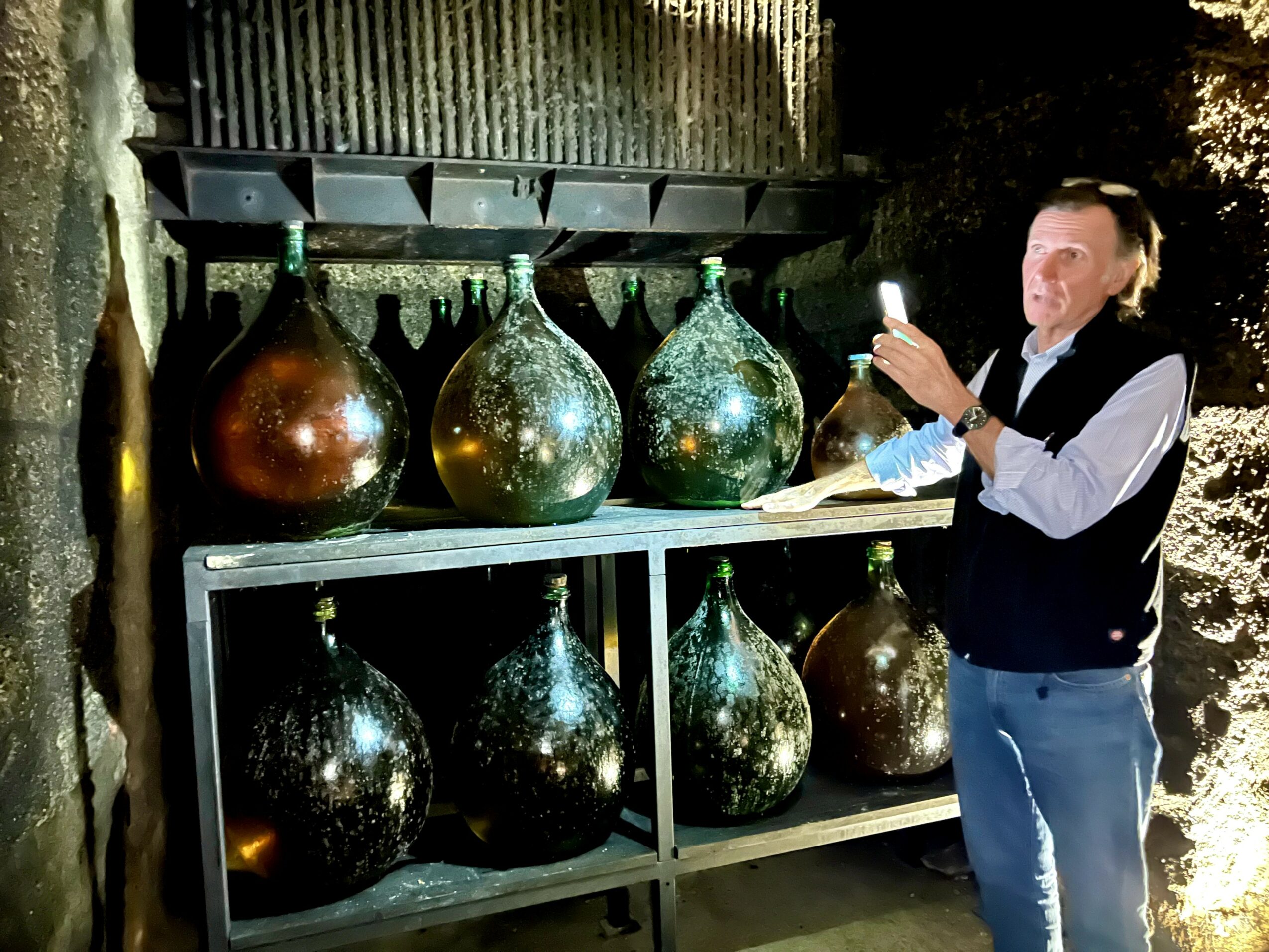 Man taking a photo with a smartphone next to large glass bottles on shelves outdoors at night.