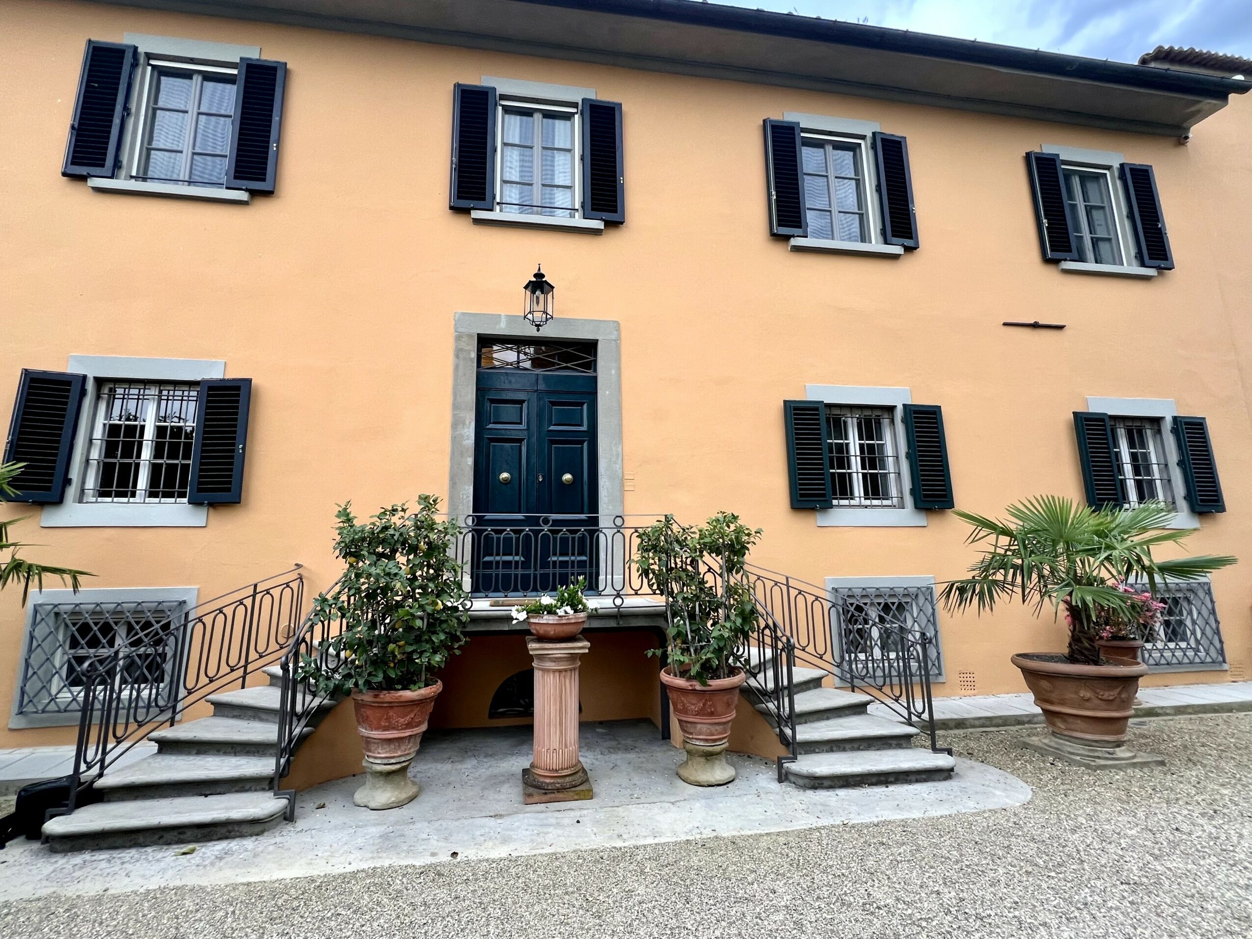 Yellow building with black shutters, blue door, and potted plants on stairs, with a gravel ground.
