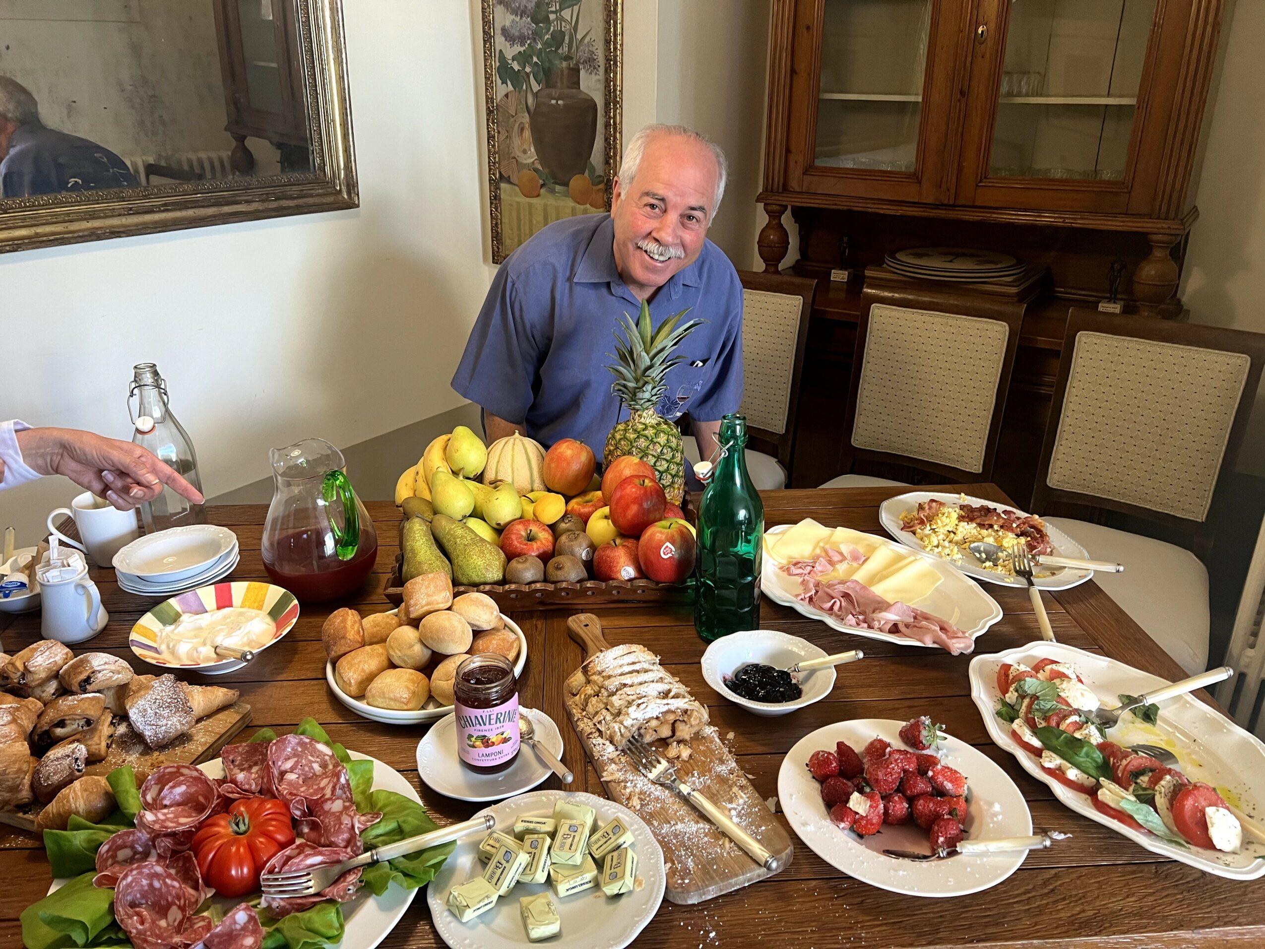 Smiling man with gray hair in blue shirt surrounded by fruits and food on a dining table.
