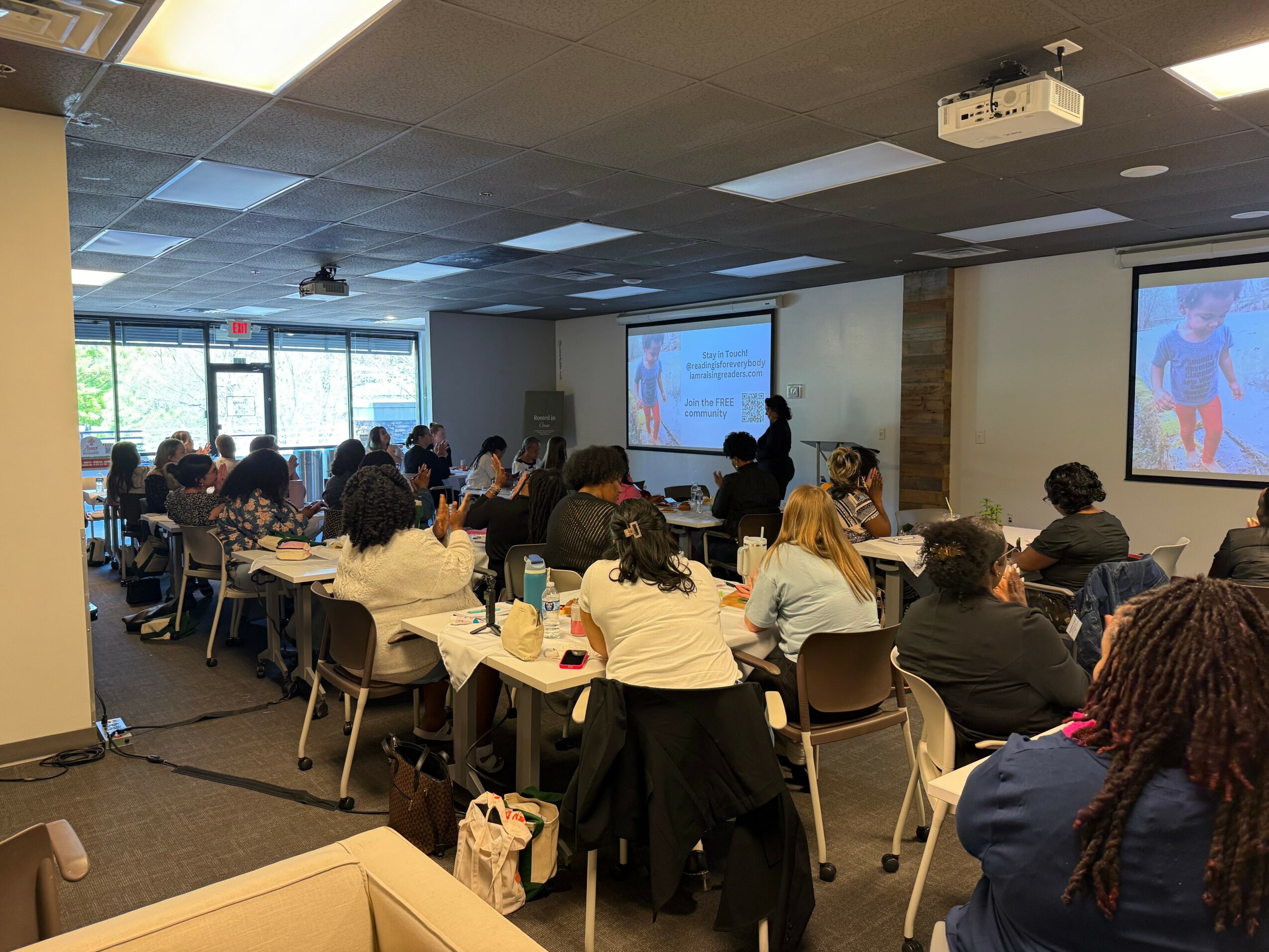 Conference room filled with people seated at tables, watching a presentation on two screens at the front.