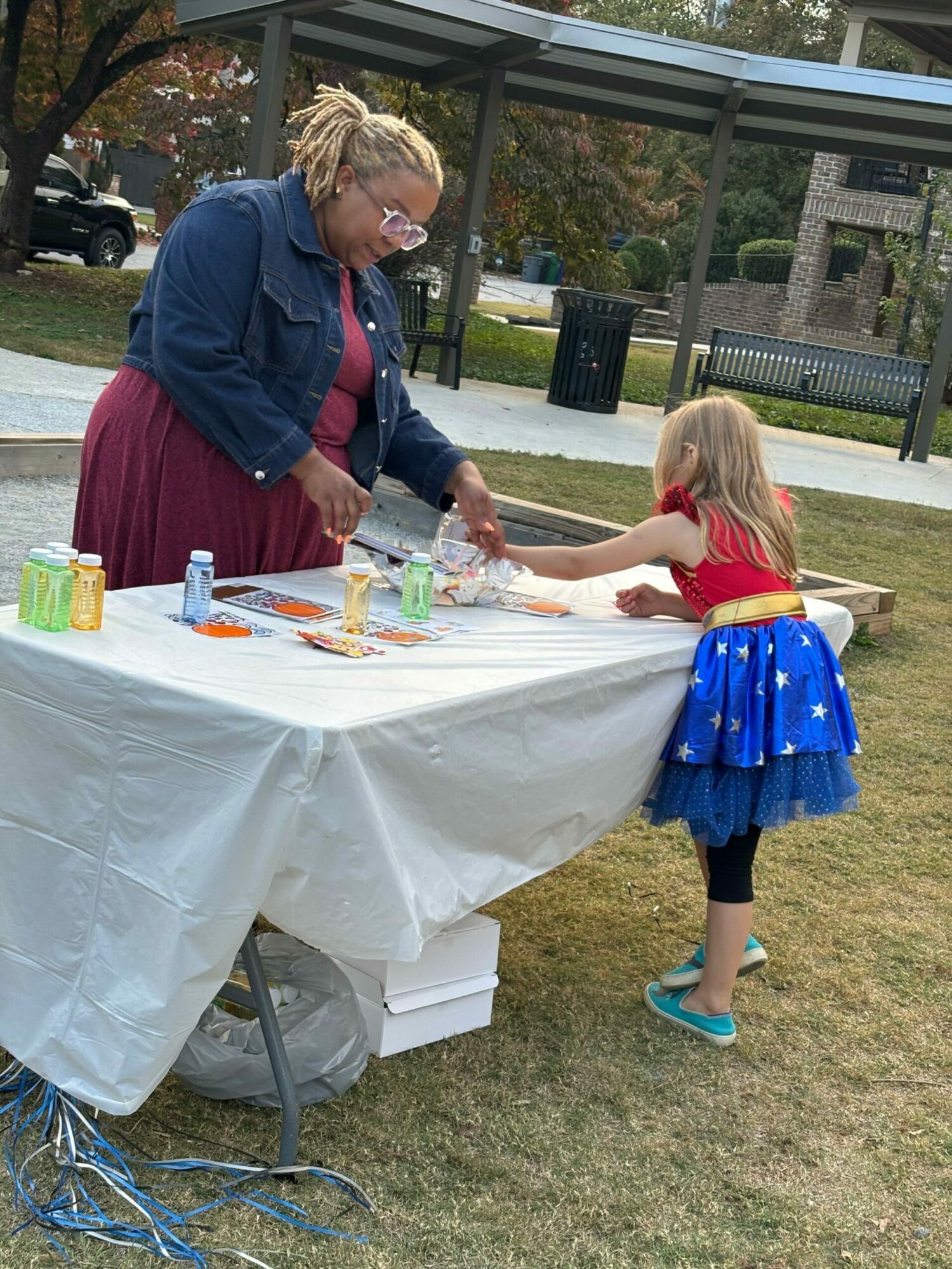 Child in a red shirt and blue skirt interacts with an adult at a table outdoors, with a grassy area and glass building in background.
