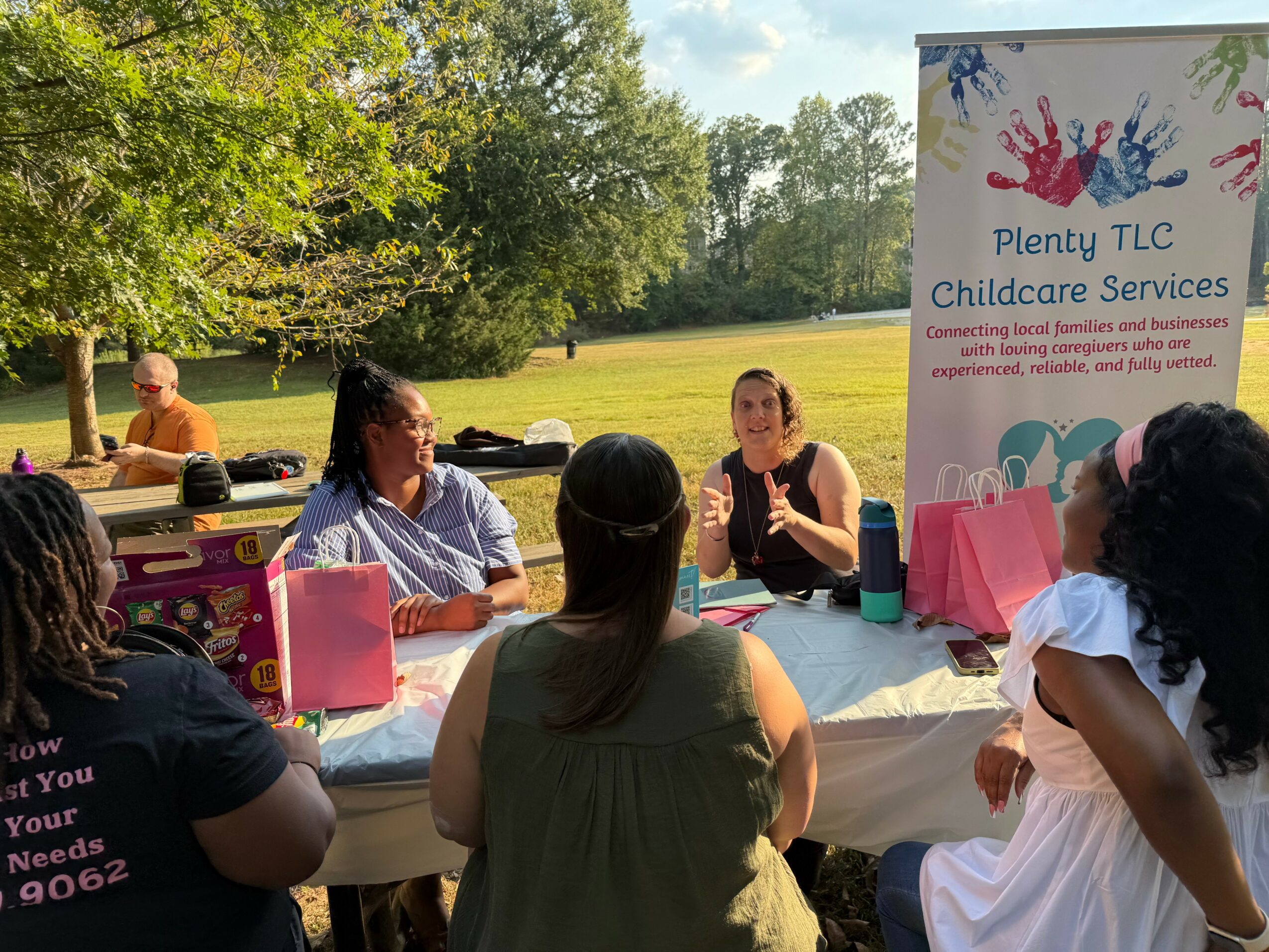 Group of women sitting at a table outdoors with a sign for childcare services in the background.