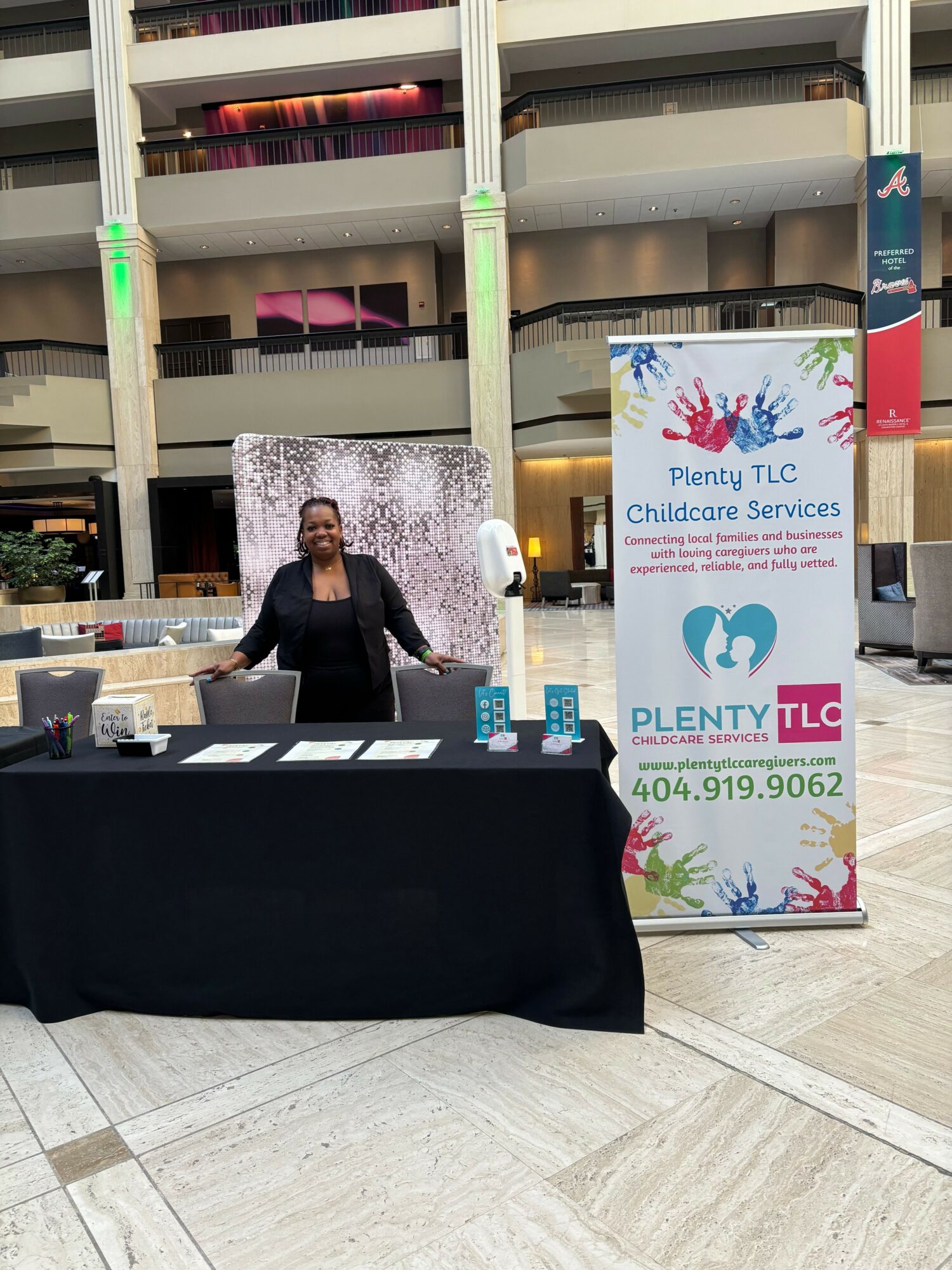 Person standing behind a table with promotional materials in a spacious indoor area, next to a colorful sign for childcare services.