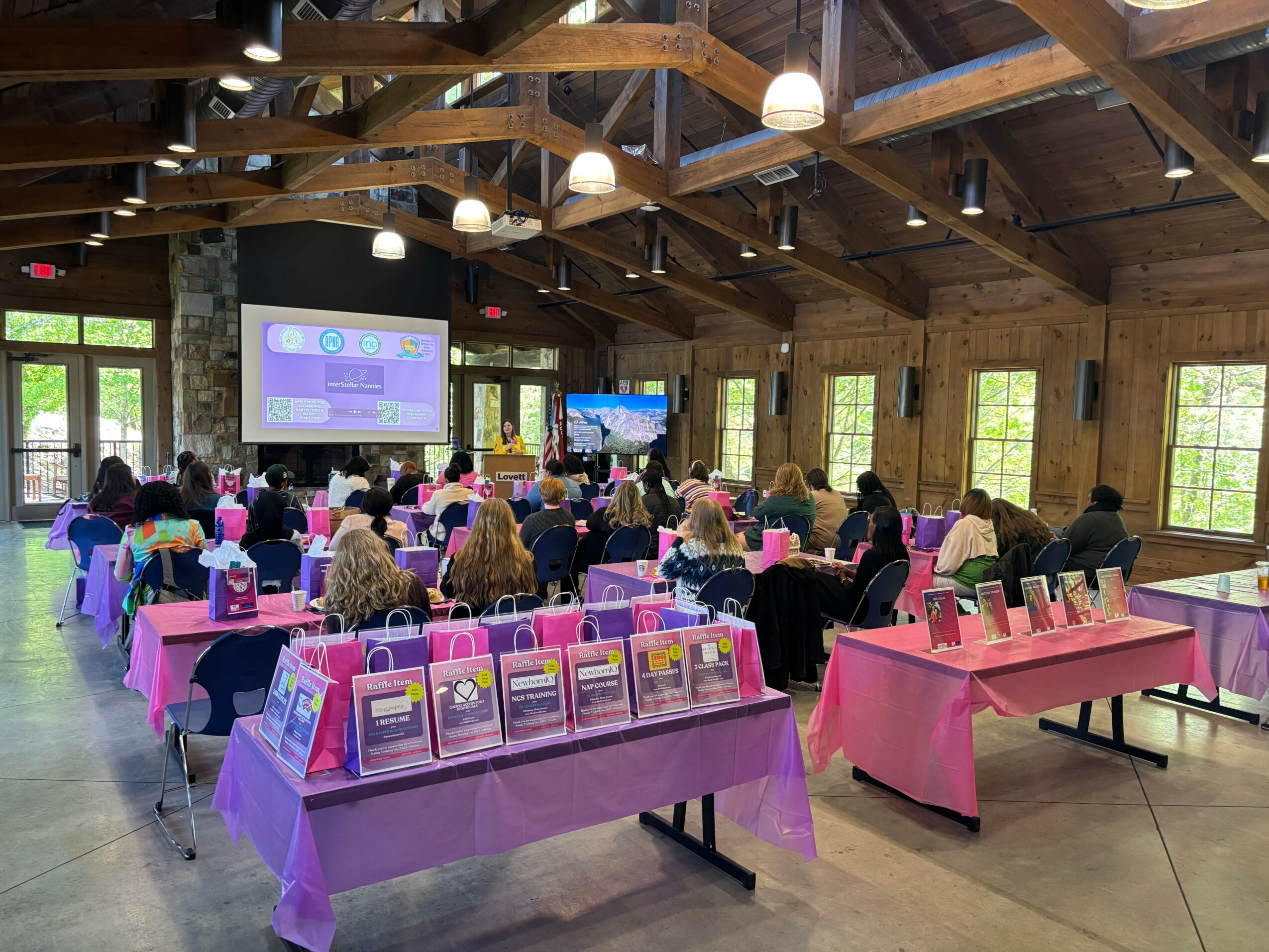 People seated at tables in a wooden hall watching a presentation on a screen, pink and purple tablecloths, windows, ceiling lights.