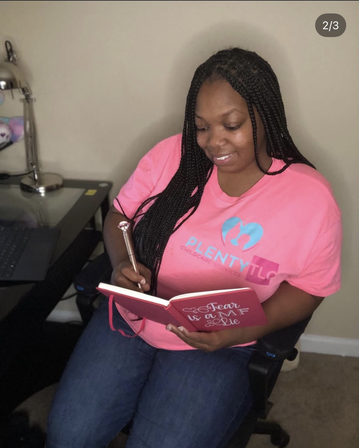 Woman with long braided hair wearing pink shirt, sitting at desk, writing in notebook.