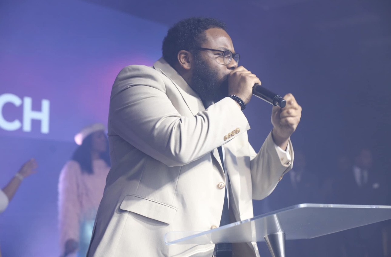 Man in white suit speaking into microphone on stage with purple background.
