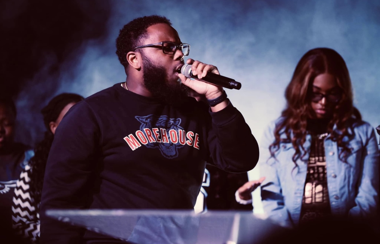 Person singing into microphone with others in background, stage lighting, dark atmosphere.