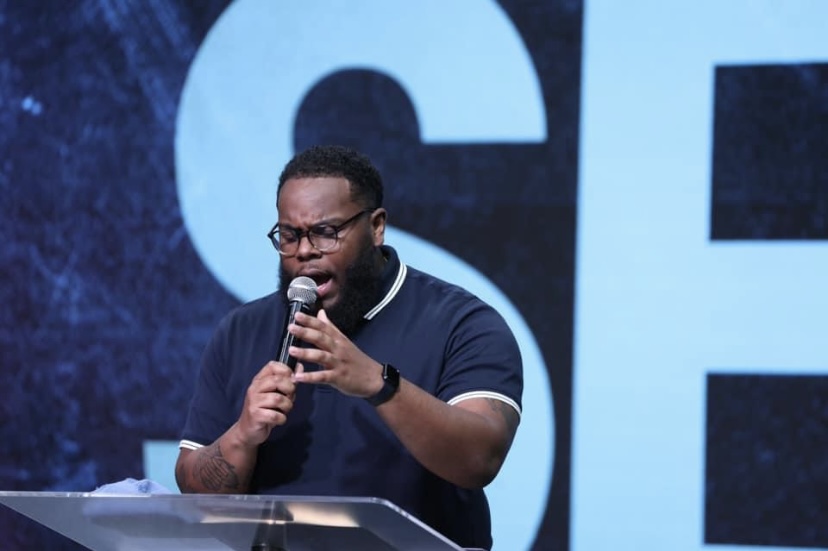 Man speaking into a microphone on stage, wearing glasses and a dark polo shirt, with a large blue background behind him.