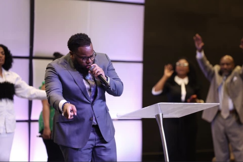 Man in a suit speaking into a microphone on stage with people raising hands behind him.