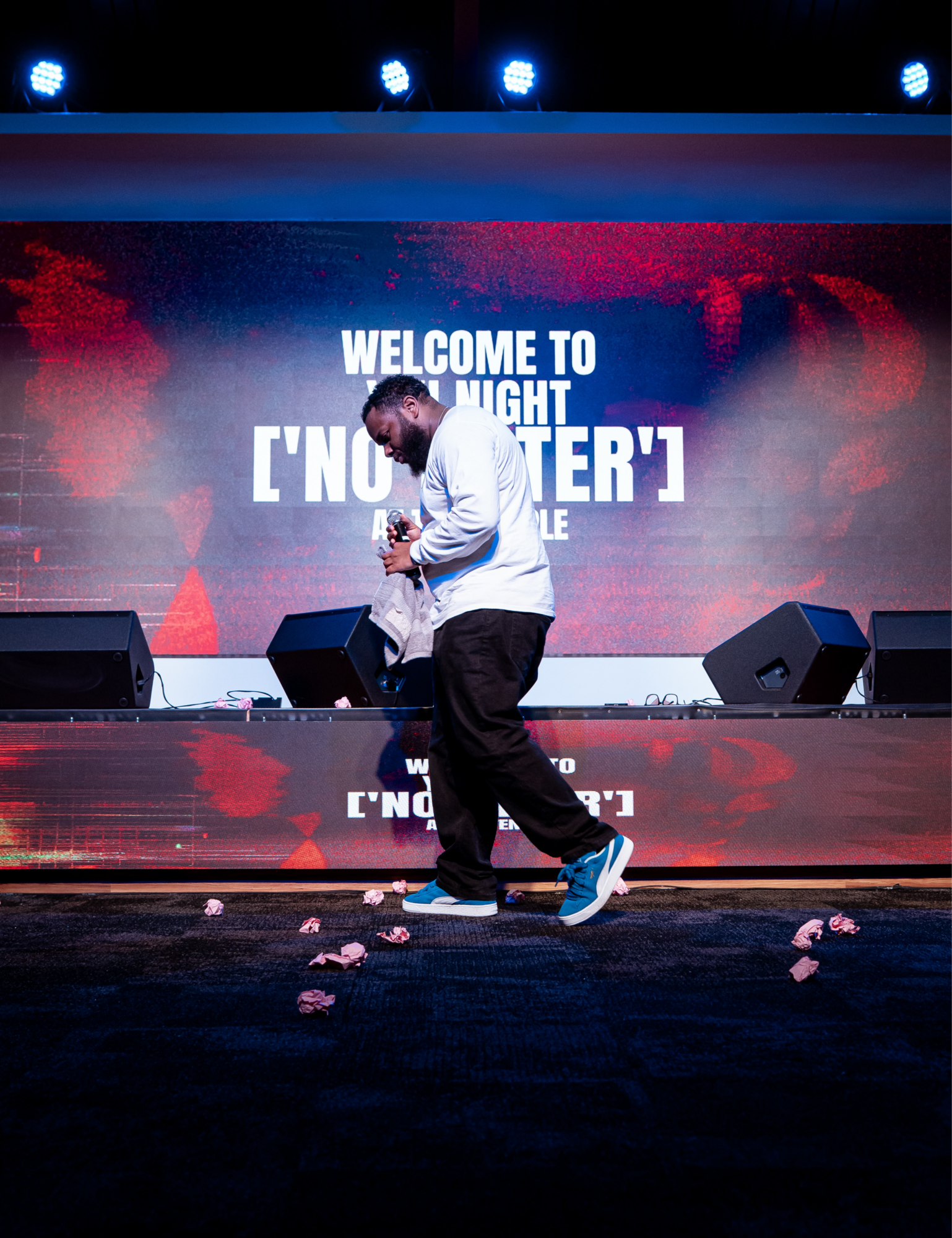 Person standing on stage with a large screen behind displaying text, stage lights above, scattered paper on floor.