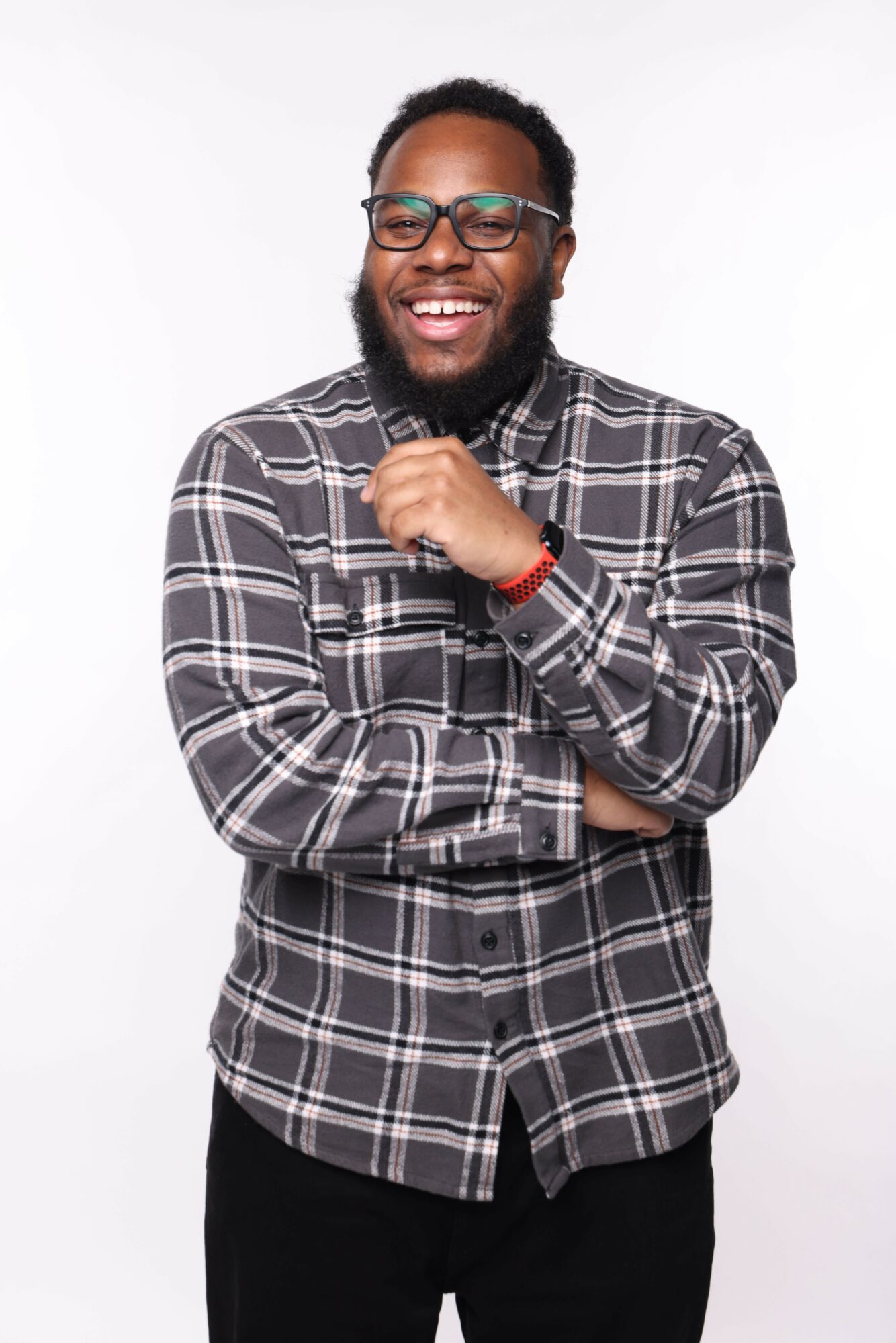 Smiling man with glasses, beard, wearing plaid shirt, standing with arms crossed against a white background.