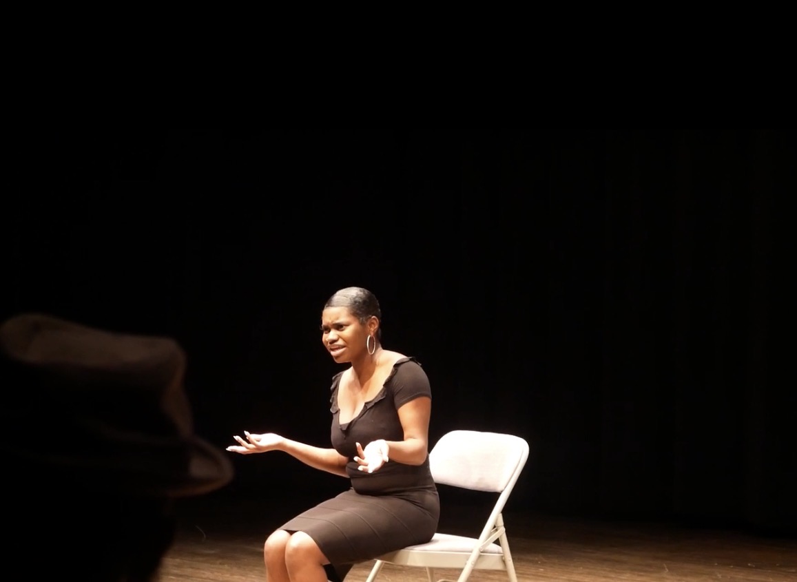 Woman sitting on a chair on stage, speaking or performing, with a dark background.
