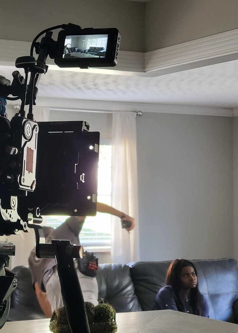 Camera on tripod capturing a woman sitting on a sofa in a room with window and curtains.