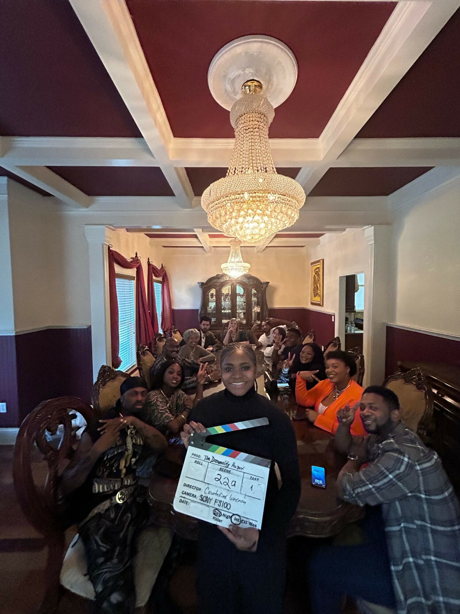 Group of people gathered around a dining table in a decorated room with a chandelier, some holding a clapperboard.