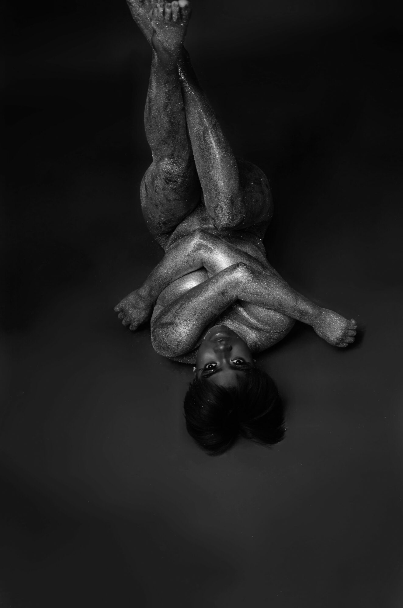 Person lying on back with legs crossed and arms over head, in a dark setting.