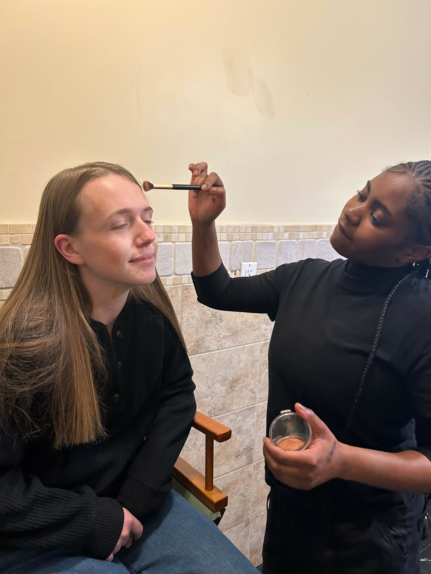 A person applying makeup to another person with closed eyes in an indoor setting.