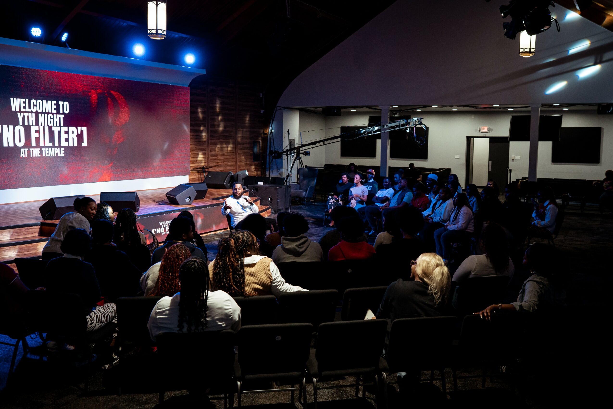 Audience seated in a dark auditorium watching a presentation on stage with a large screen displaying text.